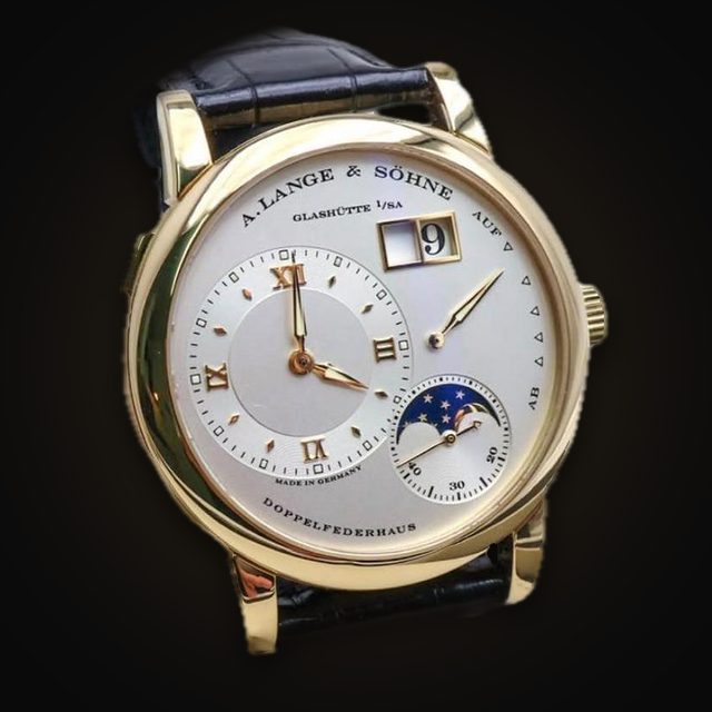 Goldene Armbanduhr mit silbernem Zifferblatt, Mondphase, Großdatum und schwarzem Lederband vor schwarzem Hintergrund